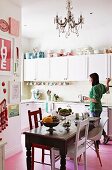 Fruit bowl on antique dining table and chairs of various colours and styles in white country-house kitchen; woman in background