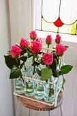 Pink roses in bottles in vintage metal bottle carrier on simple stool in front of Art Nouveau, stained-glass window