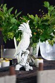 White china cockatoo ornament in front of glass vase of green leaves on rustic table
