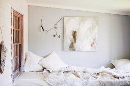 Tagesbett mit verschiedenen, weissen Plaids und Kissen in Zimmerecke vor Fenster und an hellgrau getönter Wand, modernes Bild neben Natur Fundstücken