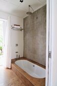 Reduziertes Badezimmer mit Betonwand und Regendusche, eingelassene weiße Badewanne mit Natursteinfliesen verkleidet