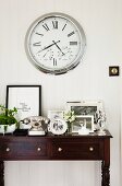 Several black and white photos in silver frames and vintage telephone on elegant console table below clock with Roman numerals on wall