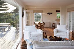 Open-plan interior of country house - armchairs with white loose covers around coffee table, dining area in background and view of furnished wooden terrace through glass wall to one side