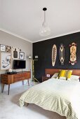 Französisches Bett mit Holz Kopfteil vor schwarz getönter Wand mit aufgehängten Skateboards, an der Seite Fifty Sideboard vor hellgrau getönter Wand mit Fernseher zwischen Zeichnungen