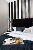 Breakfast tray on black bedspread on bed with black headboard against wall with elegant, striped black and white wallpaper