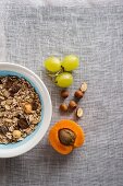 A bowl of muesli next to grapes, nuts and an apricot