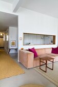 Offener Wohnraum mit Loungebereich, pastellrosa Couch und Holz Beistelltisch vor Wand mit fensterartiger Öffnung, seitlich Blick in beleuchteten Gangbereich
