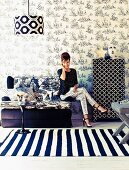 Seating area in fashionable mixture of black and white patterns; woman seated on two-seater sofa, floral wallpaper and striped rug