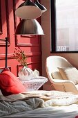 Red pillow on bed, wire mesh side table and white armchair below window against wooden panelled wall painted light red