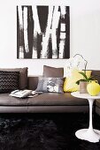 Dark grey leather couch with scatter cushions in various colours below black and white picture on wall; white orchid on white classic side table to one side