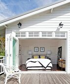 Sonnenbeschienenes weisses Holzhaus mit Terrasse und Blick durch offene Falttür in Schlafzimmer
