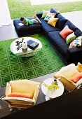 View down into spacious seating area with round glass table, green rug, modern couch and yellow, Neo-Rococo armchairs