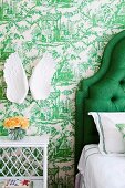 Bedside table next to partially visible bed with green, upholstered headboard; angel's-wings ornament on wall with toile de jouy wallpaper