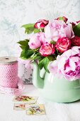 A bunch of peonies and roses in an old-fashioned green metal jug on a shelf in front of a wallpapered wall