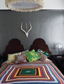 Bunte gehäkelte Tagesdecke und Kissen auf Doppelbett mit geschwungenen Kopfteilen aus Holz, vor grau getönter Wand mit Tiertrophäe