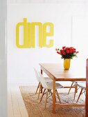 Dining area - vase of dahlias on wooden table, white classic shell chairs on sisal rug and bright yellow letters painted on wall
