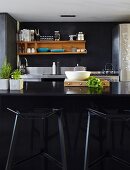 Black, designer bar stools at black counter; wooden shelves on dark wall in background