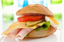 A bread roll filled with ham, cheese, tomatoes, egg and gherkins on a chopping board