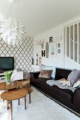 Designer artichoke lamp and set of retro side tables in front of black leather sofa; wallpaper with Oriental tile motif and vintage ornaments in background