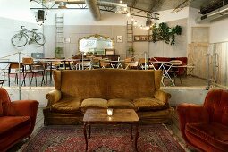 Velvet-covered couch and armchairs in large loft apartment; dining tables on platform and gilt-framed mirror on tiled wall in background