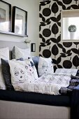 Scatter cushions with comic-style covers on bed; black and white floral wallpaper on one wall in background