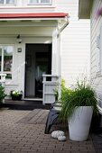 Weisses Blumengefäss mit Pflanze auf Terrasse, im Hintergrund Veranda mit offener Tür vor weißem Holzhaus