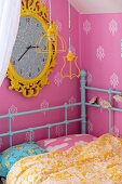 Kinderbett mit türkisem Retro Metallgestell in Zimmerecke, grosse Wanduhr mit gelbem, verziertem Rahmen auf rosa Tapete mit Ornamentmuster