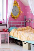Child's bed with turquoise, retro metal frame below clock with yellow, ornate frame on pink wallpaper