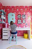 Girl sitting at desk in nostalgic bedroom; pink wallpaper with floral pattern