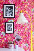 Stehleuchte mit gelbem Gestell und nostalgischem Stoffschirm, im Hintergrund gerahmte Fotos an tapezierter Wand mit farbigem Blumenmuster