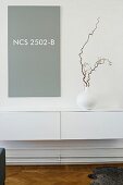 Twig in vase on white, minimalist floating sideboard below grey panel with NCS colour code on wall