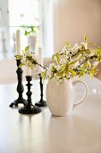 Branches of white blossom in china jug and candlesticks on white table