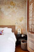 Bed with white bed linen, bedside table and laundry basket in front of wall with vintage patina