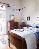 Bed with patchwork bedspread, bunting above wooden chest of drawers and white wood-panelling in cheerful, country-house, child's bedroom
