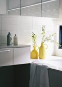 White wall tiles and sprigs of flowers in yellow retro vases and edge of bathtub in bathroom