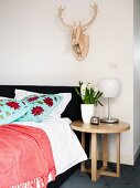 Round oak side table next to upholstered bed with floral scatter cushions below stylised antlers on wall