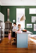 Mother and children in open-plan kitchen with little girl sitting on counter and stainless steel cooker against dark green wall in background