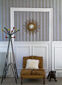 Fellkissen auf braunem Sessel und Retro Garderobenständer vor Wand mit weisser, halbhoher Holzverkleidung, darüber Streifentapete