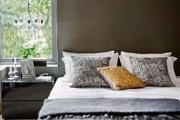 Mirrored bedside cabinet below window next to modern double bed with scatter cushions against grey wall