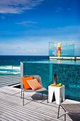 Holiday atmosphere - cane chairs and white stools on wooden deck, pool with transparent walls and view of Pacific Ocean in background