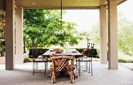 Sitzplatz mit rustikalem Stuhl aus Ästen und filigranen Metallstühlen auf grosszügiger, überdachter Terrasse mit Gartenblick