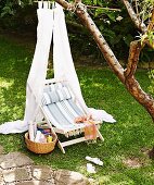 Ländlicher Liegestuhl mit gestreiftem Bezug unter weißem Stoffbaldachin an Baum aufgehängt, im Garten