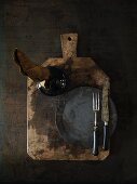 A vase, a plate and cutlery on a rustic wooden board on a dark surface