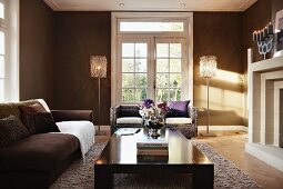 Glass double doors flanked by standard lamps in living room painted brown with elegant seating area in front of fireplace