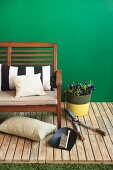 Potted plant next to garden bench and various gardening tools arranged on wooden decking against green-painted wall