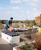 Sitzbank mit gestreiften Polstern, Tisch und Stühle auf Dachterrasse; Mann richtet Sitzkissen an