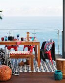 Holzbank, gedeckter Tisch und rote Retrostühle auf Terrasse mit Meerblick durch transparente Glasbrüstung