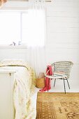 Vintage Rattanstuhl neben Bett mit geblümten Plaid vor weisser Holzwand und Fenster mit luftigem Vorhang