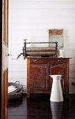 White metal water jug on dark wooden floor in front of old mangle on top of antique wooden cabinet