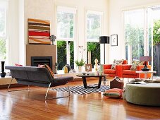 Seating in various colours, coffee table and open fireplace in corner of high-ceilinged, bright interior